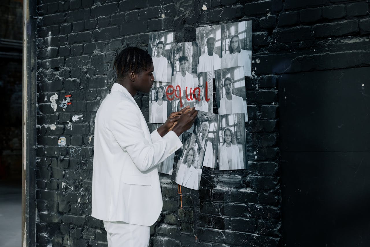 Man in white suit writing 'equal' on black and white photos on a brick wall, emphasizing equality.