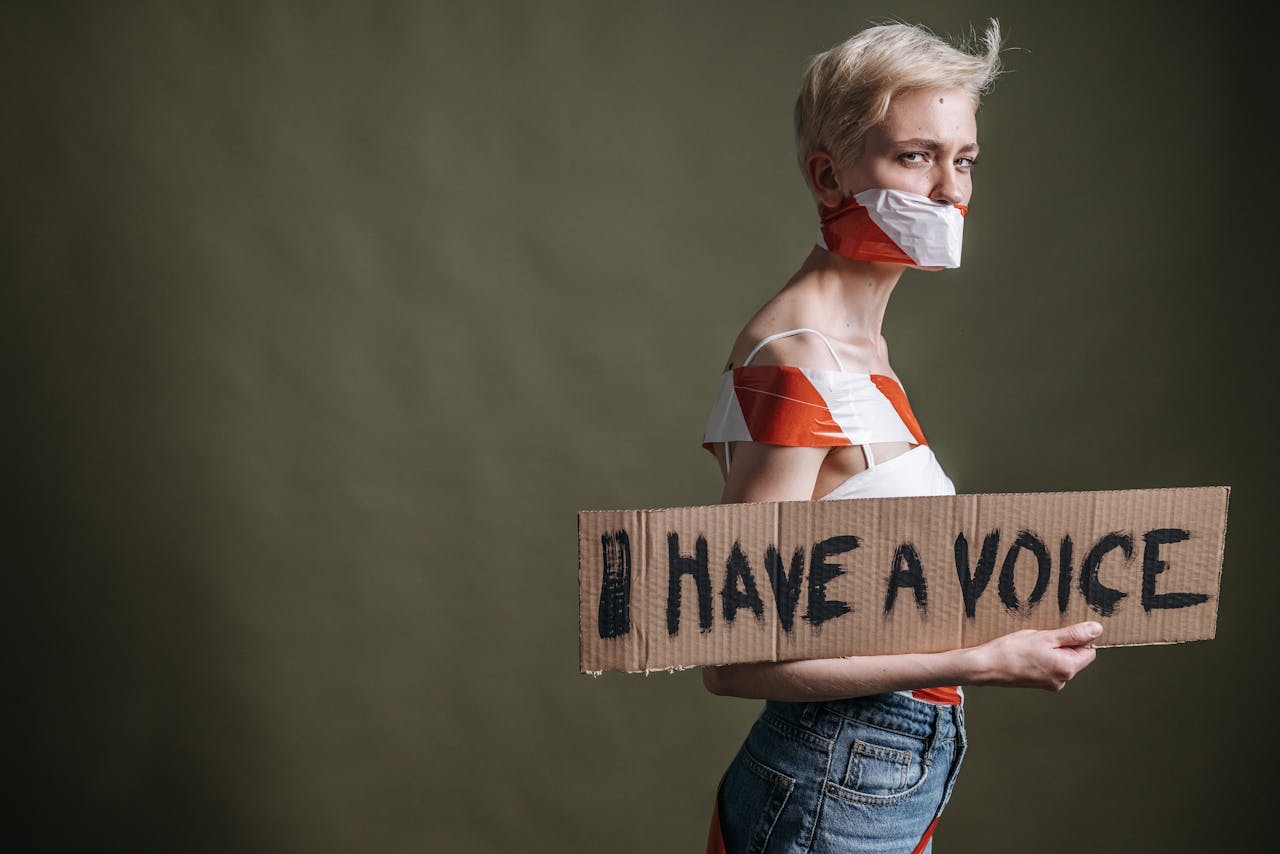 A woman wrapped in tape holds a protest placard saying 'I Have a Voice'.